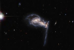 Three galaxies, the center one is barely visible and bleeds into the others on the right and left. The one on the right has a long trail of stars stretching toward the bottom. The left one still appears as an intact spiral.. A bright diffracted foreground star at far-left.